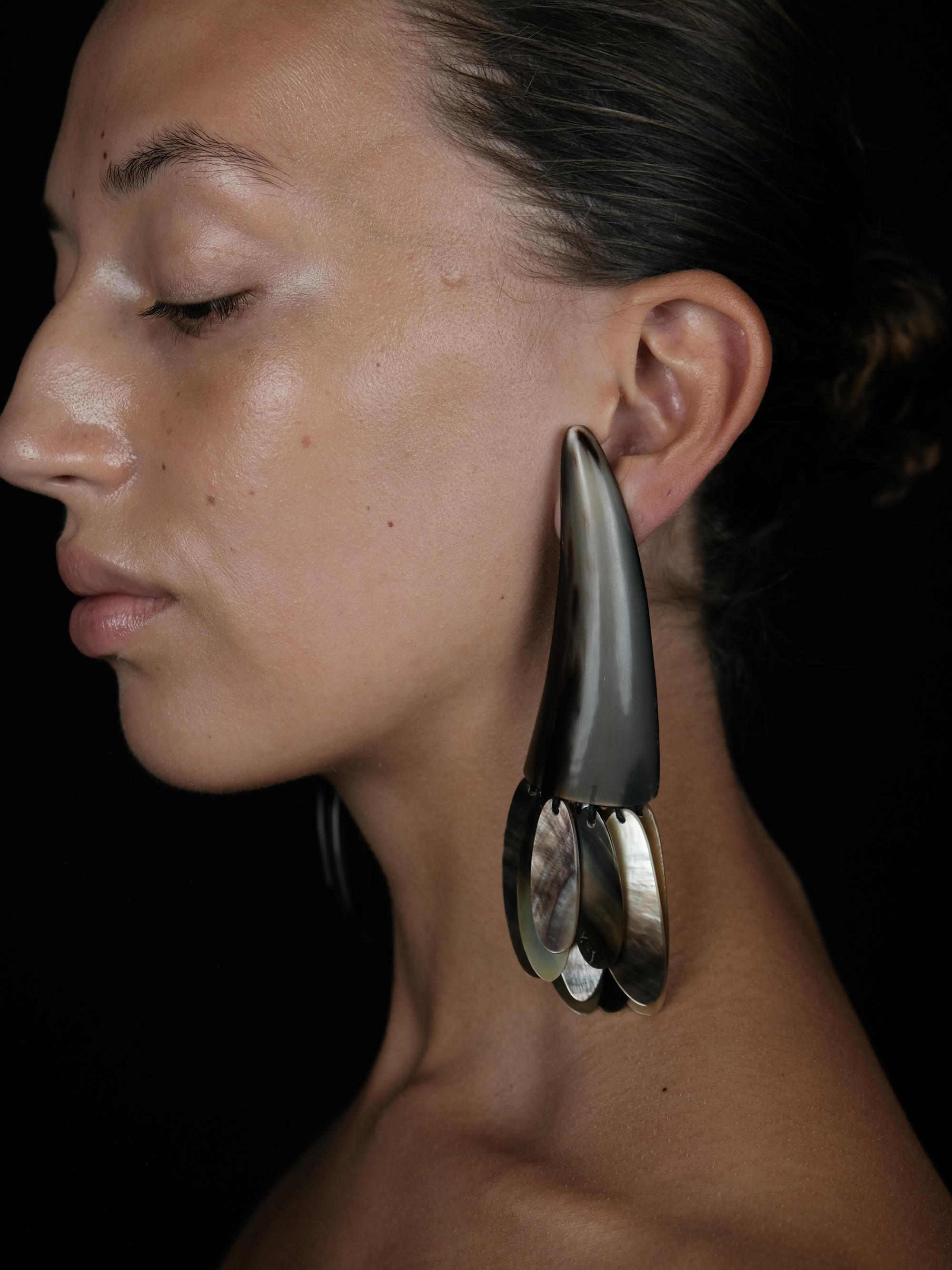 A close-up side profile of a woman wearing a dramatic, large statement earring. The earring features a dark, teardrop-shaped top section with a brushed metallic finish, hanging from a polished silver-toned hoop.