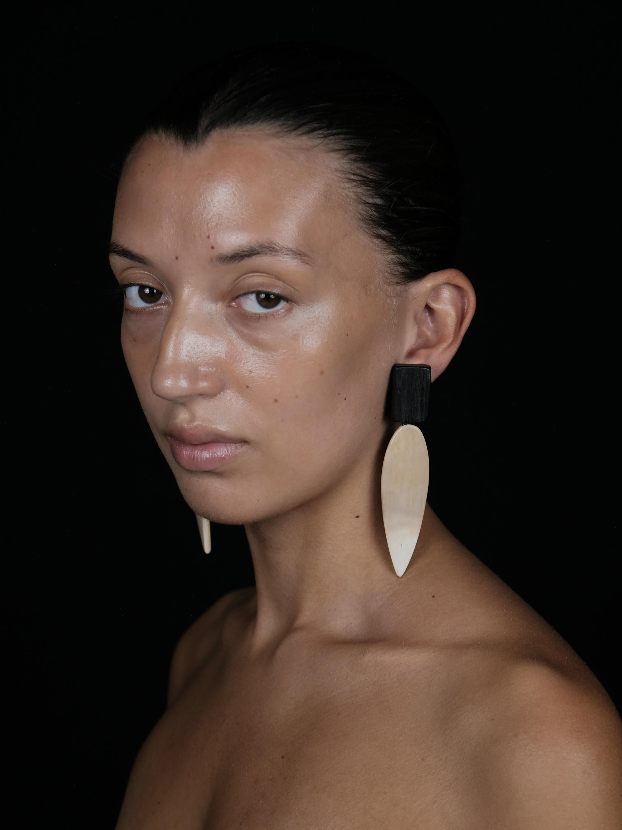 A close-up portrait of a woman with dark hair pulled back, wearing a large asymmetrical statement earring. The earring features a matte black top section and a large, teardrop-shaped natural bone or shell pendant in a warm beige tone.