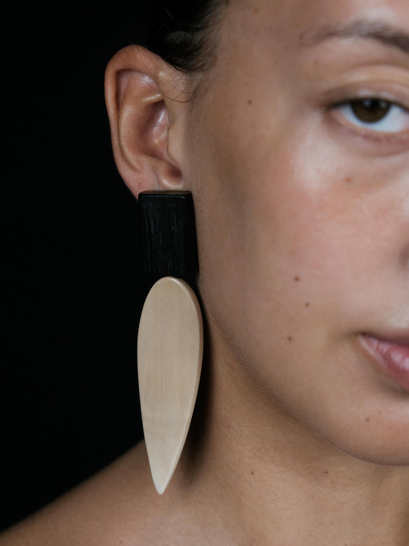 A close-up of a person's ear wearing a large statement earring with a matte black top section and a natural light wood teardrop-shaped drop.