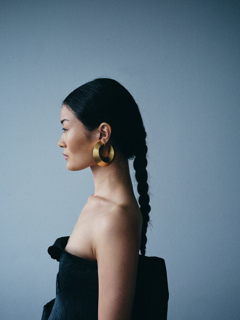 A woman in profile wearing large, chunky gold hoop earrings and a black strapless top, set against a solid slate gray background.