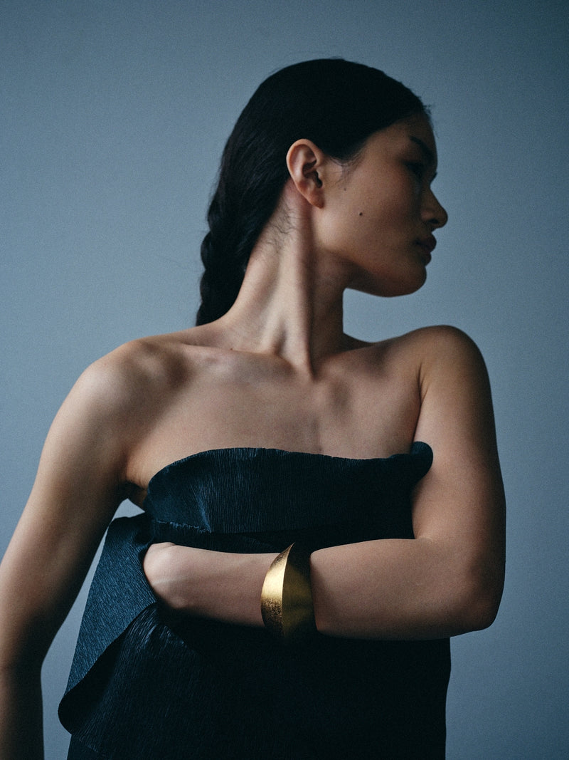 A woman in profile wearing a dark strapless top and a wide gold cuff bracelet, photographed against a muted blue-gray studio background.