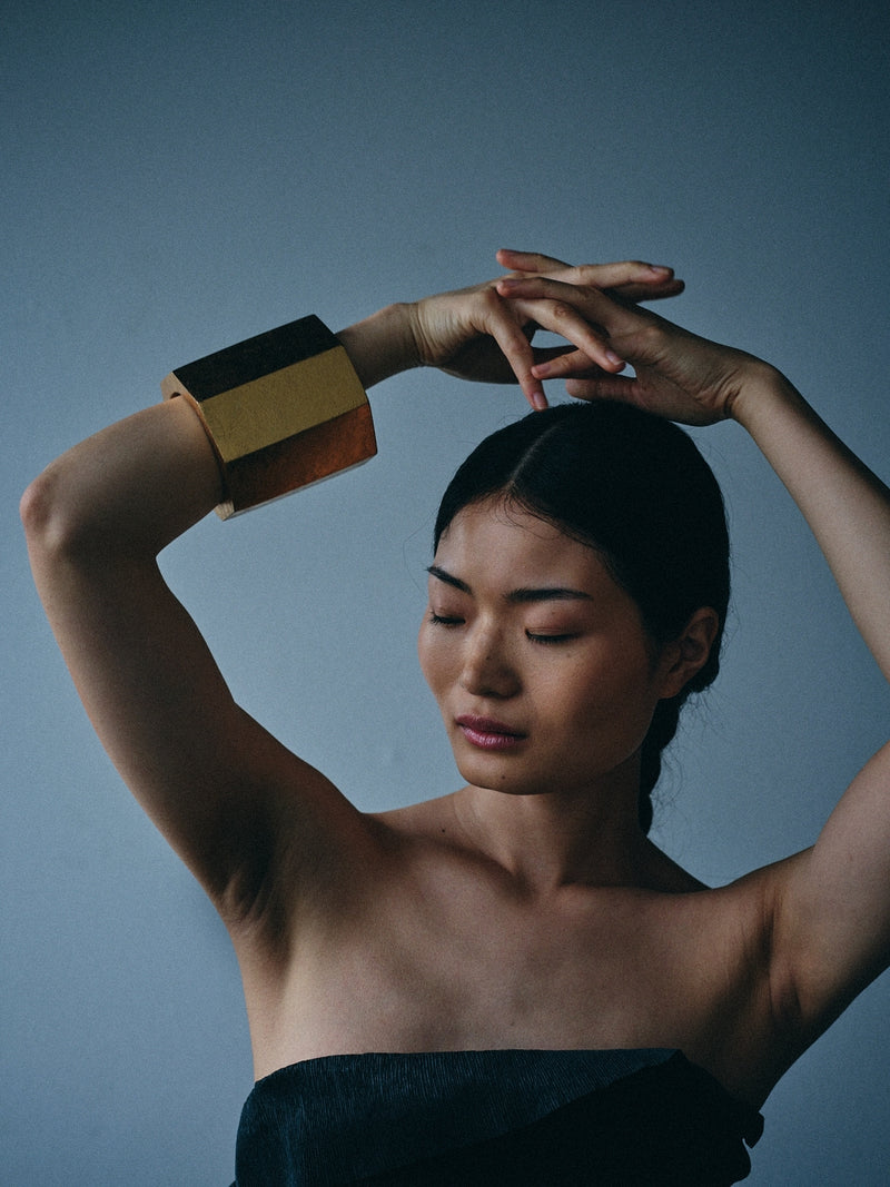 A woman with her hair pulled back wears a wide, chunky gold and black cuff bracelet on her raised arm, posing against a muted slate blue background.
