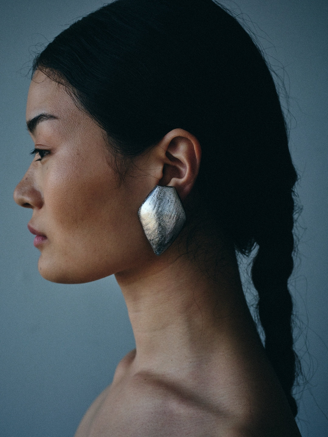 A close-up side profile of a woman wearing a large, organic-shaped metallic silver earring against a dark gray background.
