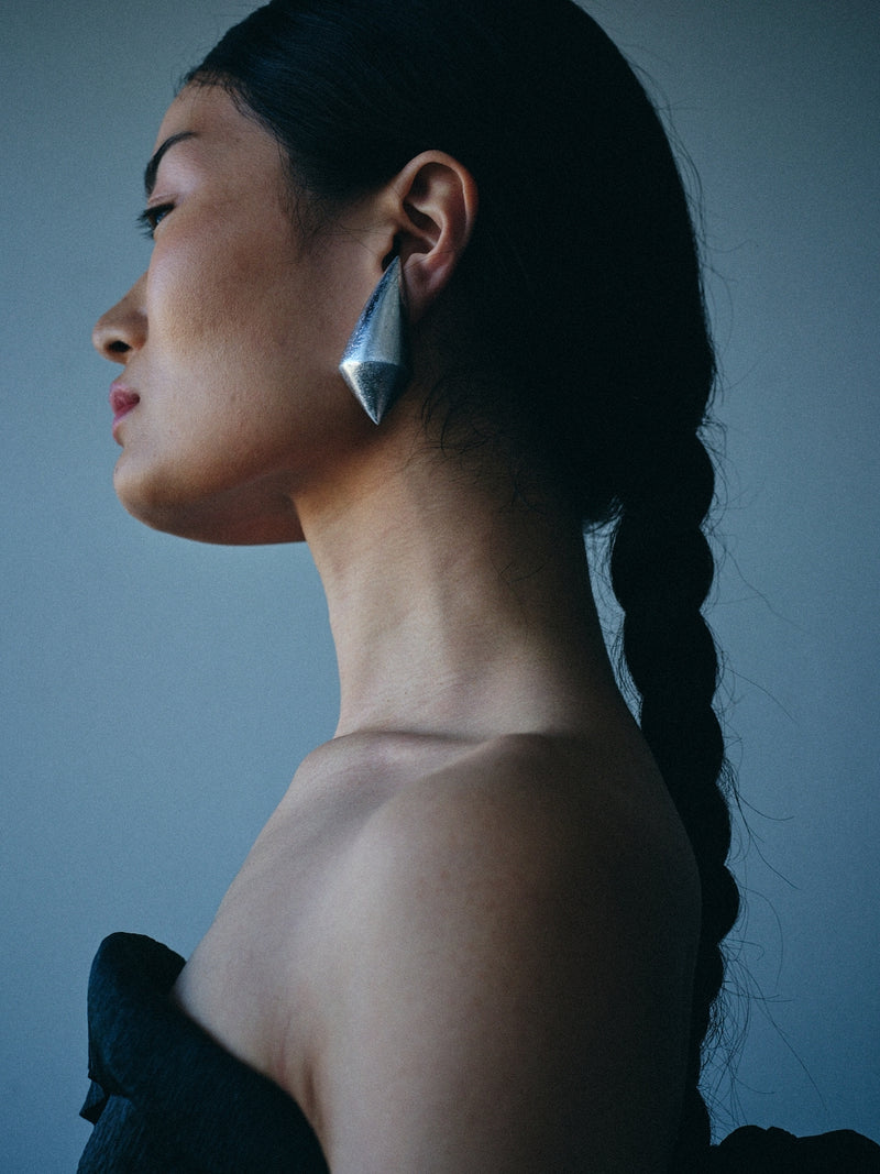 A side-profile portrait of a woman with dark hair in a braid, wearing a large, faceted silver geometric earring against a muted blue-gray background.