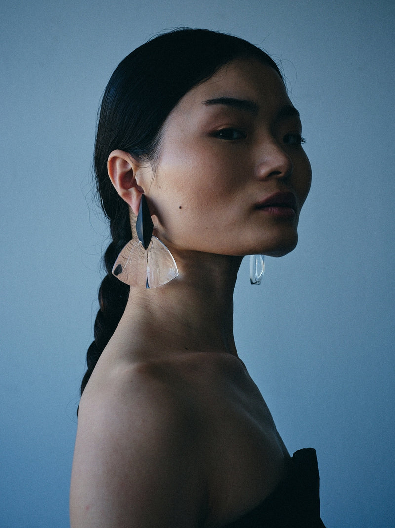 A woman in profile wearing large, asymmetrical statement earrings made of layered translucent beige and dark gray materials, set against a solid blue-gray background.