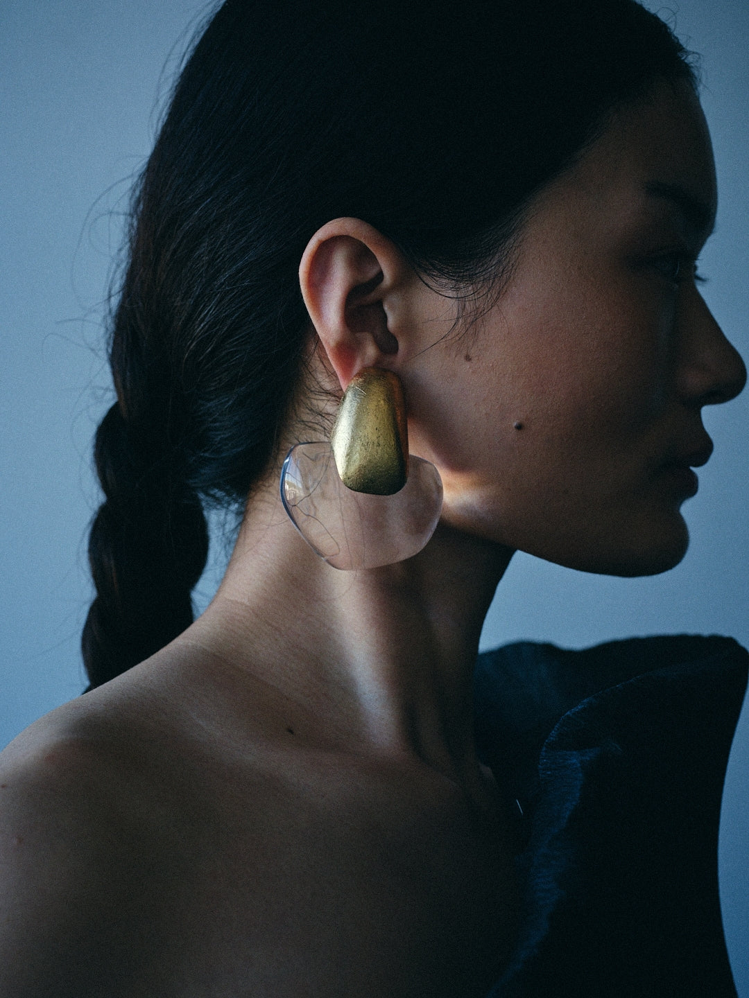A close-up side profile of a woman wearing a large, sculptural statement earring. The earring features a textured, metallic gold top section and a clear, organic-shaped lower section.
