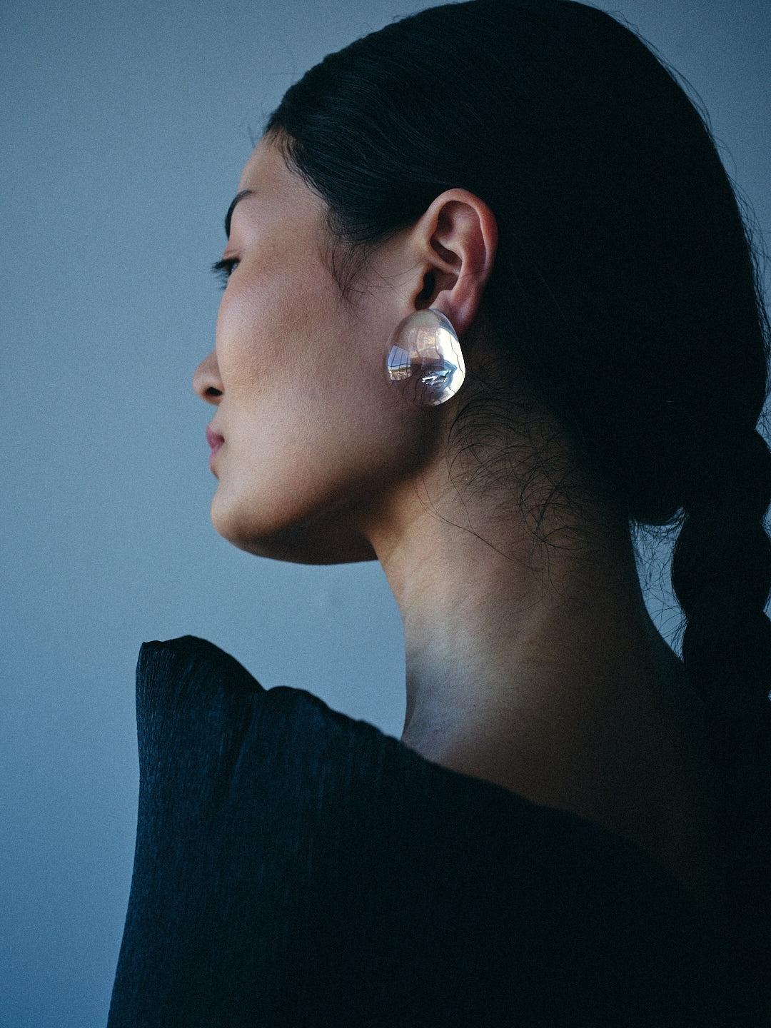 A side-profile portrait of a woman with dark hair tied back, wearing a large, clear, faceted teardrop-shaped earring against a muted blue-gray background.
