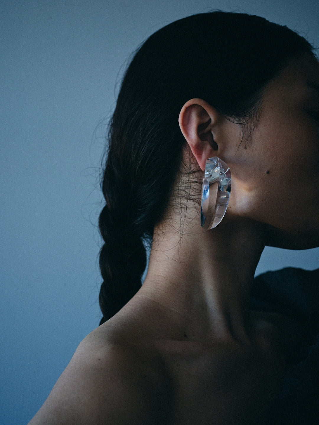 A close-up side profile of a woman with dark hair in a braid, wearing a large, chunky clear acrylic hoop earring against a muted blue-gray background.