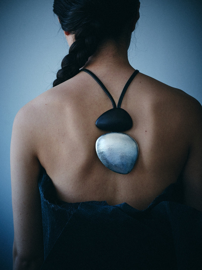 A woman shown from behind wearing a unique black cord pendant with two smooth pieces, one black and one silver, resting on her upper back.