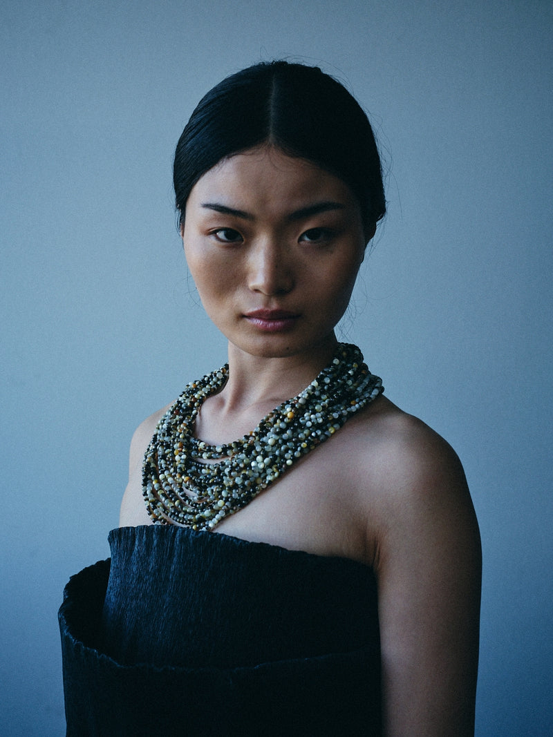 A model with dark hair pulled back wears a multi-strand beaded pearl necklace with metallic gold, silver, and green beads, paired with a dark strapless top against a cool blue-gray background.