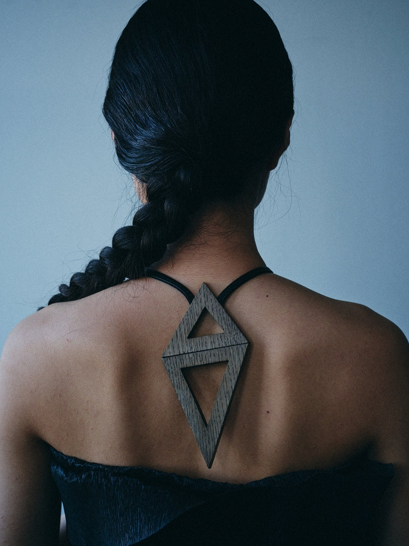 A woman viewed from behind wearing a dark wood geometric back ornament with black straps, set against a cool blue-gray background.