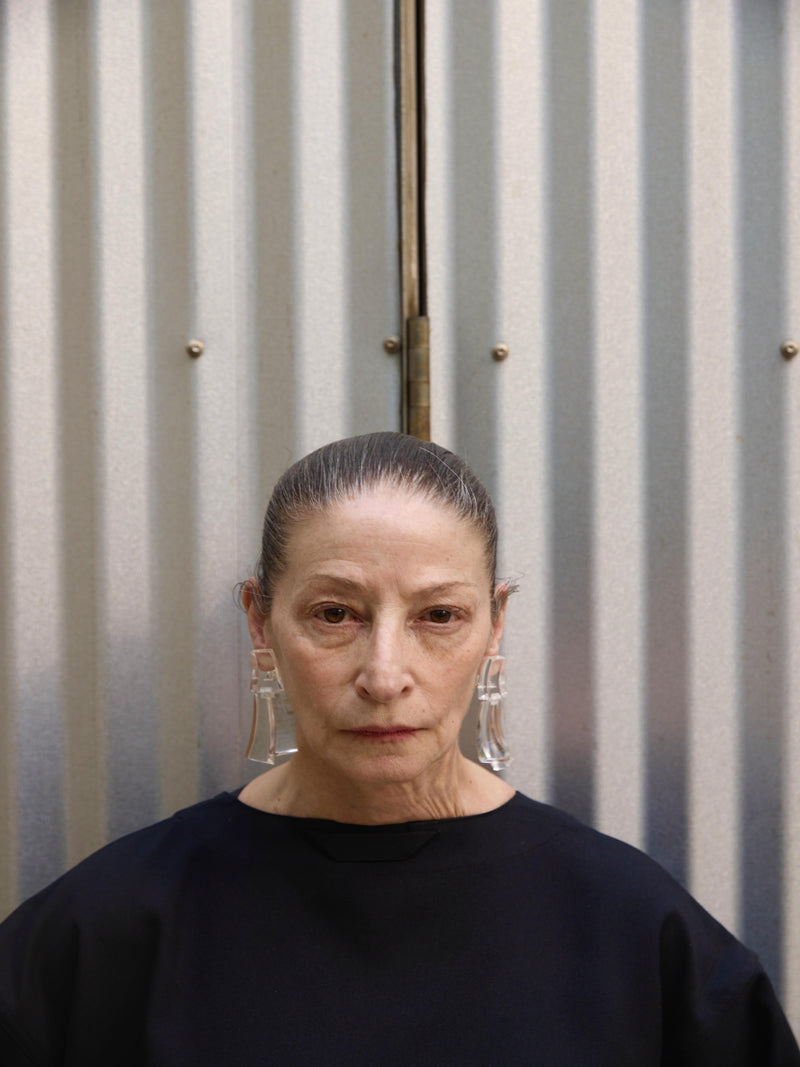 Person wearing a black top and transparent earrings against a corrugated metal background