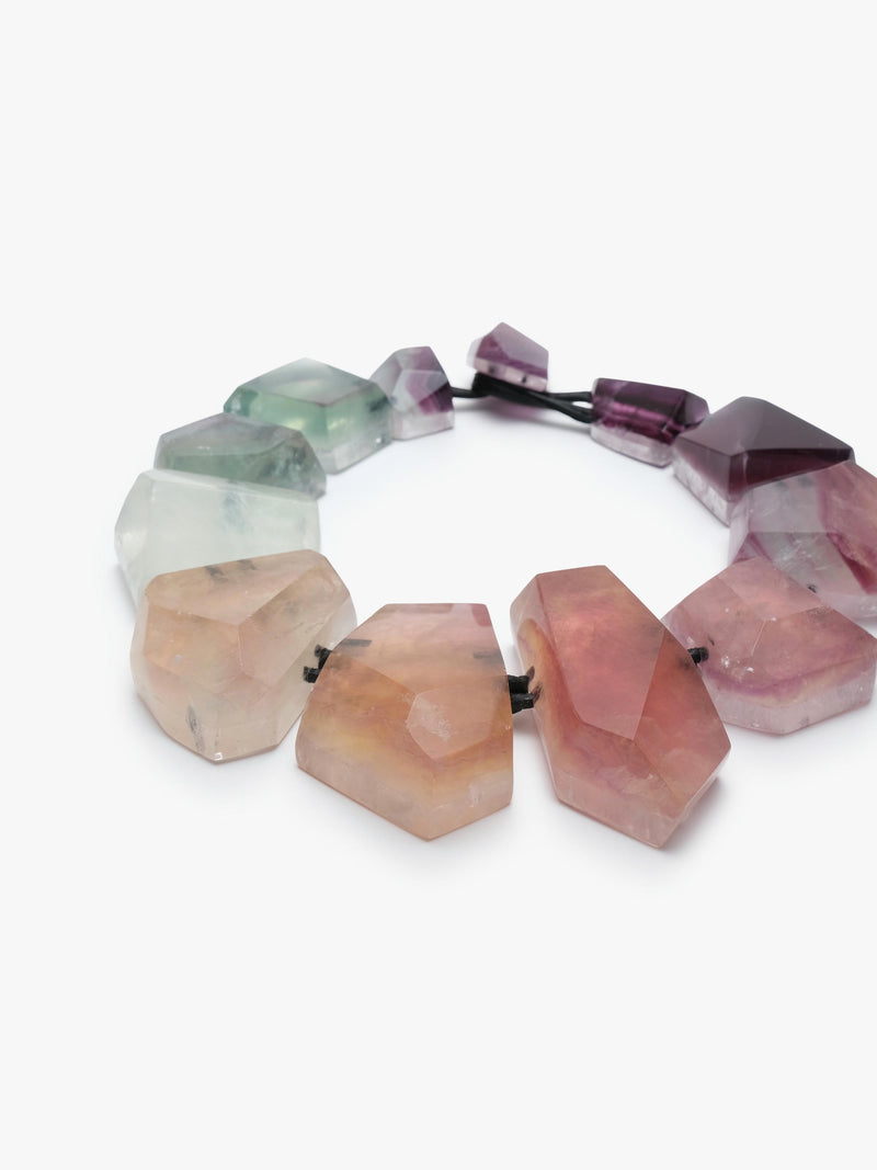 A necklace made of irregular, faceted gemstone beads in a gradient of colors from pale green and white to pink and deep purple, strung on a black elastic band, shown against a plain white background.