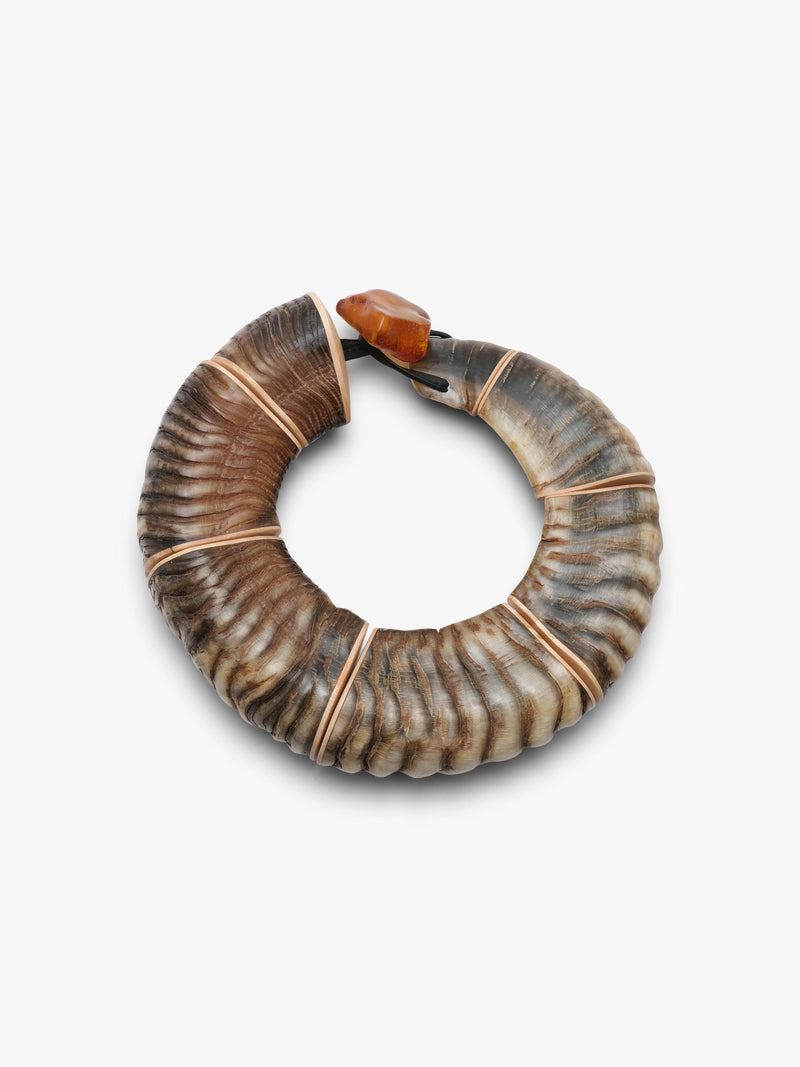 A handmade circular necklace crafted from a large rams horn, featuring natural brown and beige ridges, bound with a leather strand, and secured with a polished amber toggle clasp, shown on a light gray background.