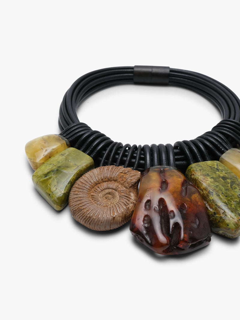 A close up of a black leather necklace, with natural stone beads in brown, green and yellow colours, displayed on a white background.