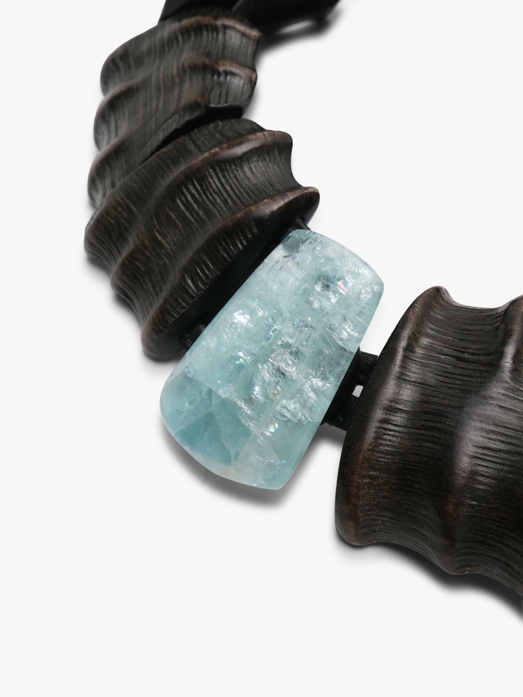 A close-up of a statement necklace featuring a textured dark brown wood-like segmented collar and a central light blue faceted crystal bead, set against a plain white background.