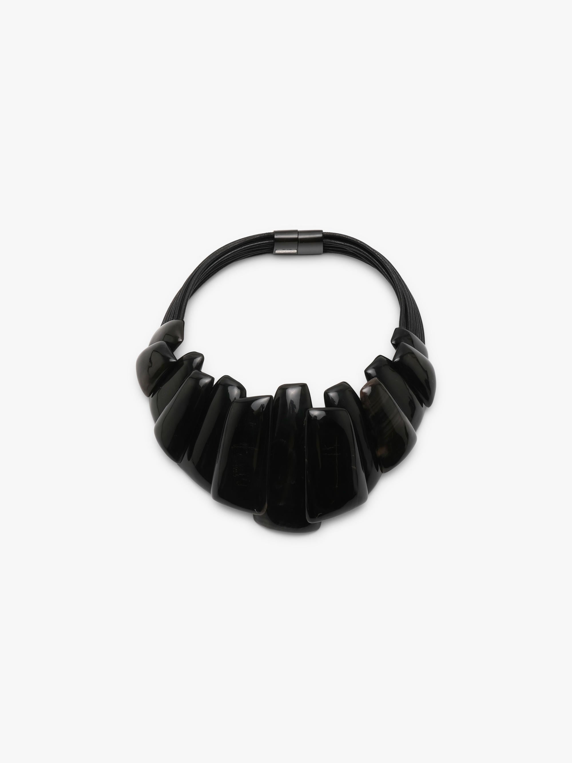 A black statement collar necklace with a sculptural, layered ruffled design and a small metallock at the top, shown on a plain light gray background.