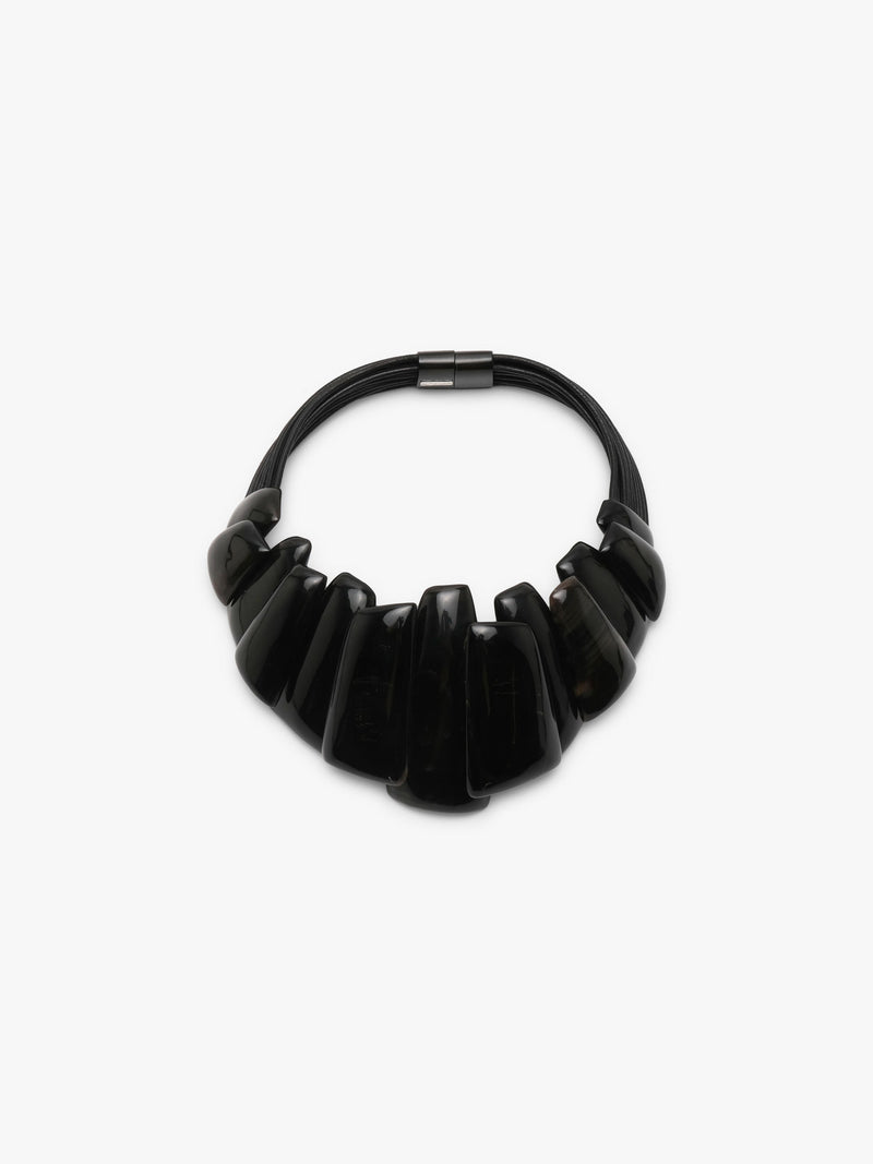 A black statement collar necklace with a sculptural, layered ruffled design and a small metallock at the top, shown on a plain light gray background.
