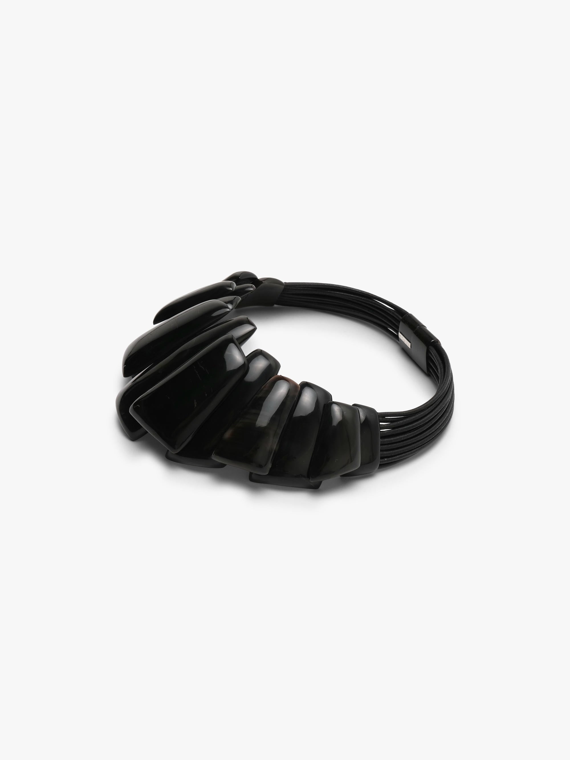 A black statement collar necklace with a sculptural, layered ruffled design and a small metallock at the top, shown on a plain light gray background.