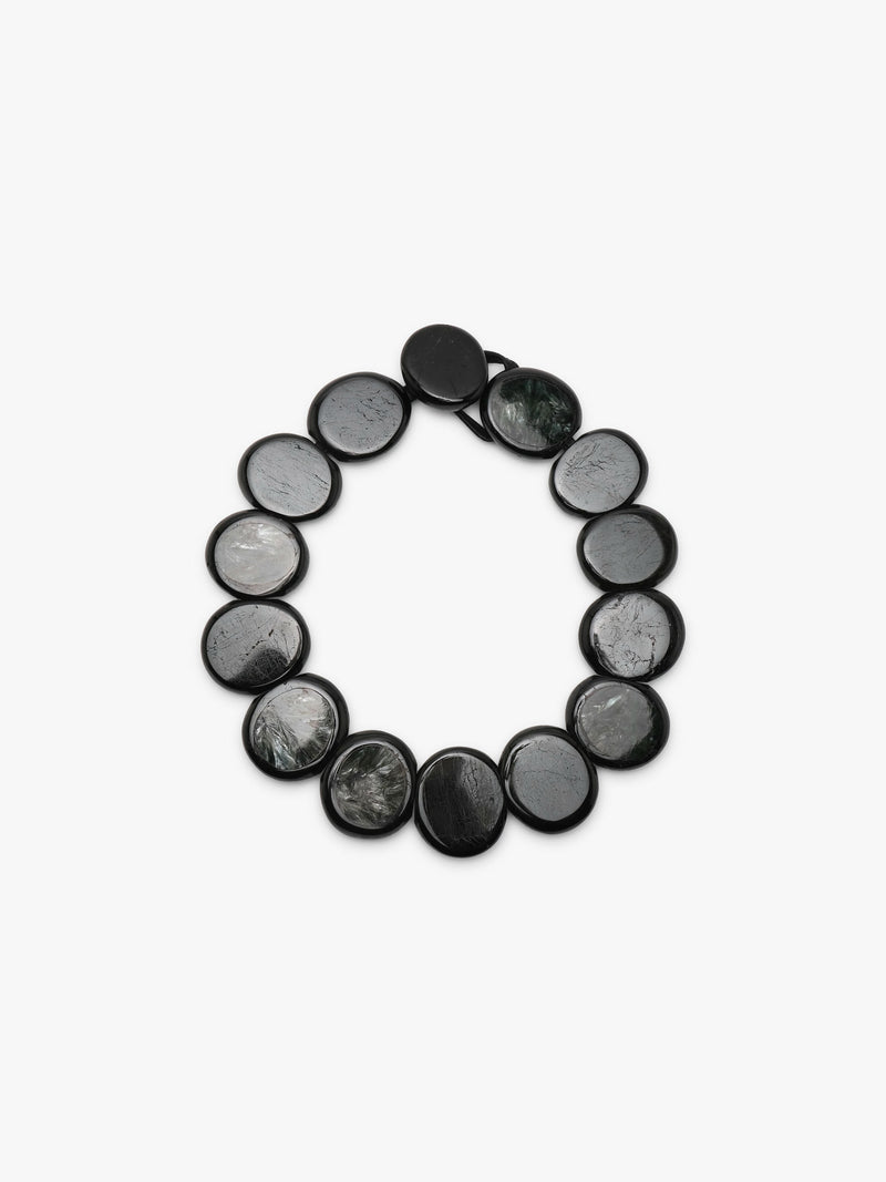 A beaded necklace made of oval dark gray to black stone beads with natural marbled patterns, arranged in a circle on a light gray background.