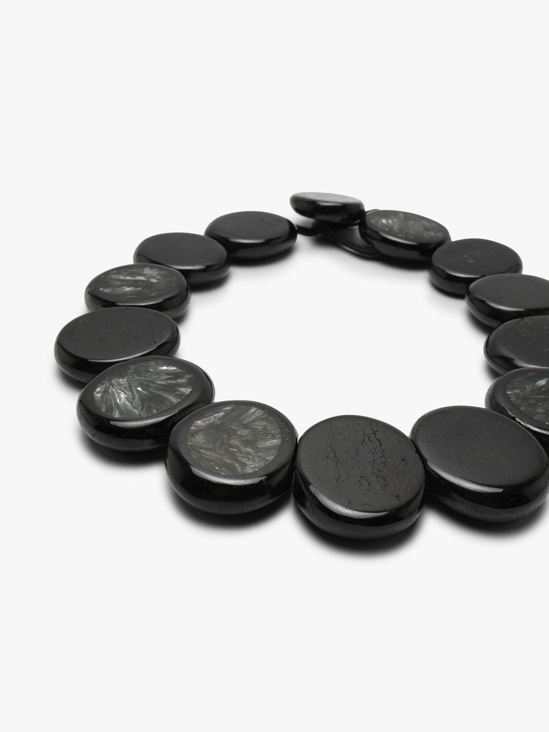 A beaded necklace made of oval dark gray to black stone beads with natural marbled patterns, arranged in a circle on a light gray background.