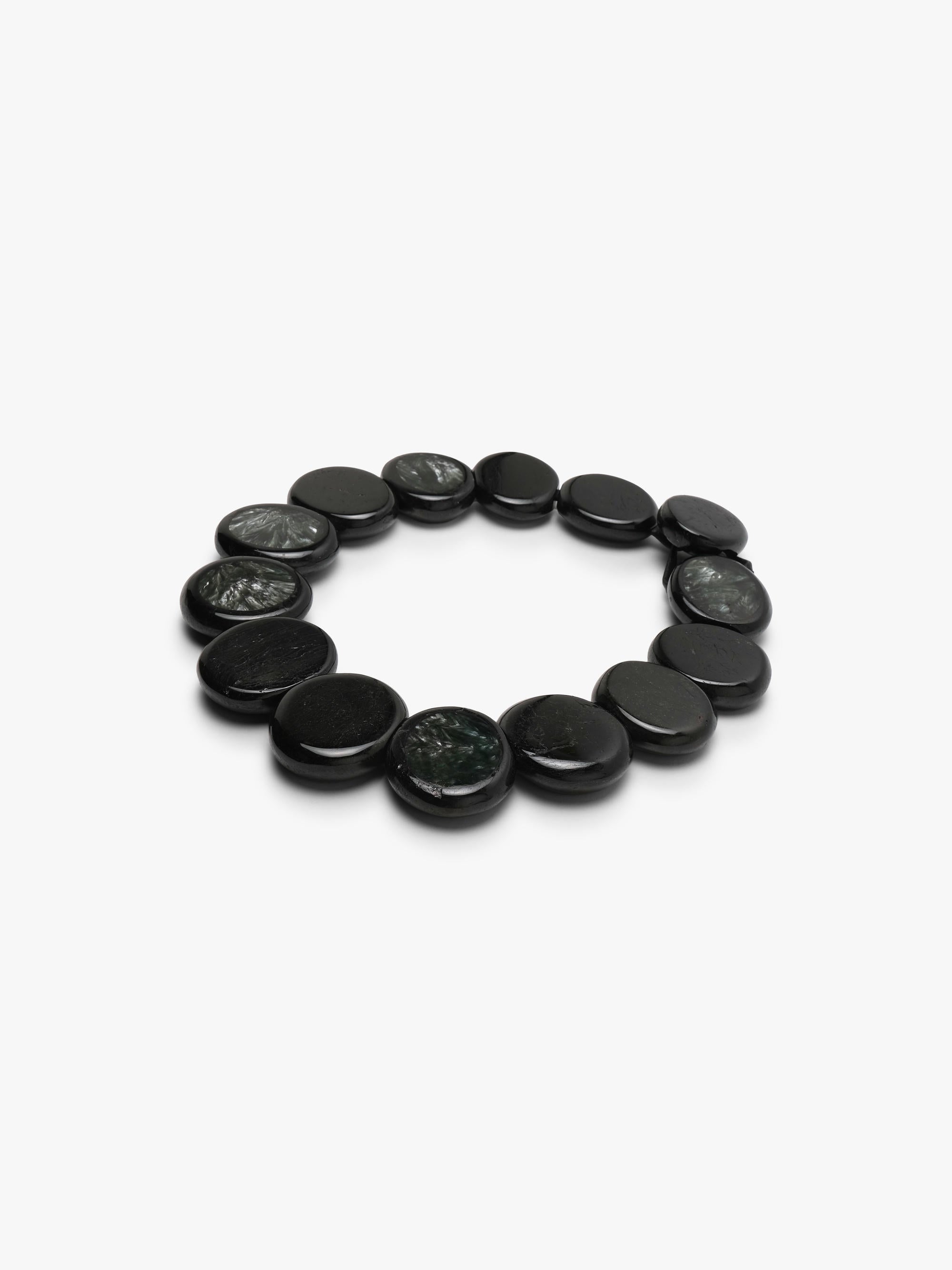 A beaded necklace made of oval dark gray to black stone beads with natural marbled patterns, arranged in a circle on a light gray background.