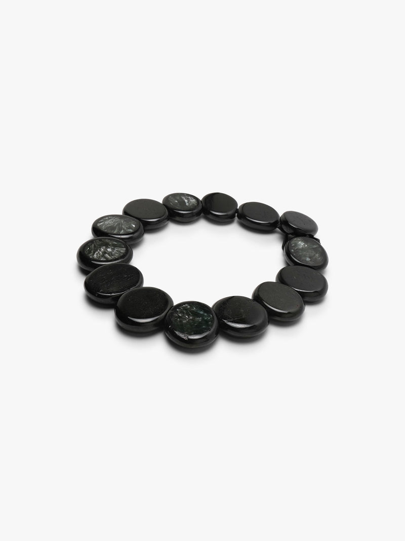 A beaded necklace made of oval dark gray to black stone beads with natural marbled patterns, arranged in a circle on a light gray background.
