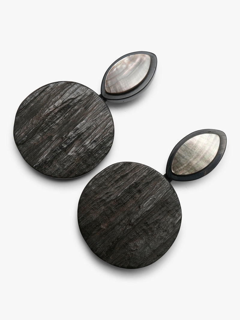 A pair of statement earrings featuring large, circular dark gray horn-grain discs connected to smaller, teardrop-shaped studs with iridescent mother-of-pearl centers, set against a clean white background.