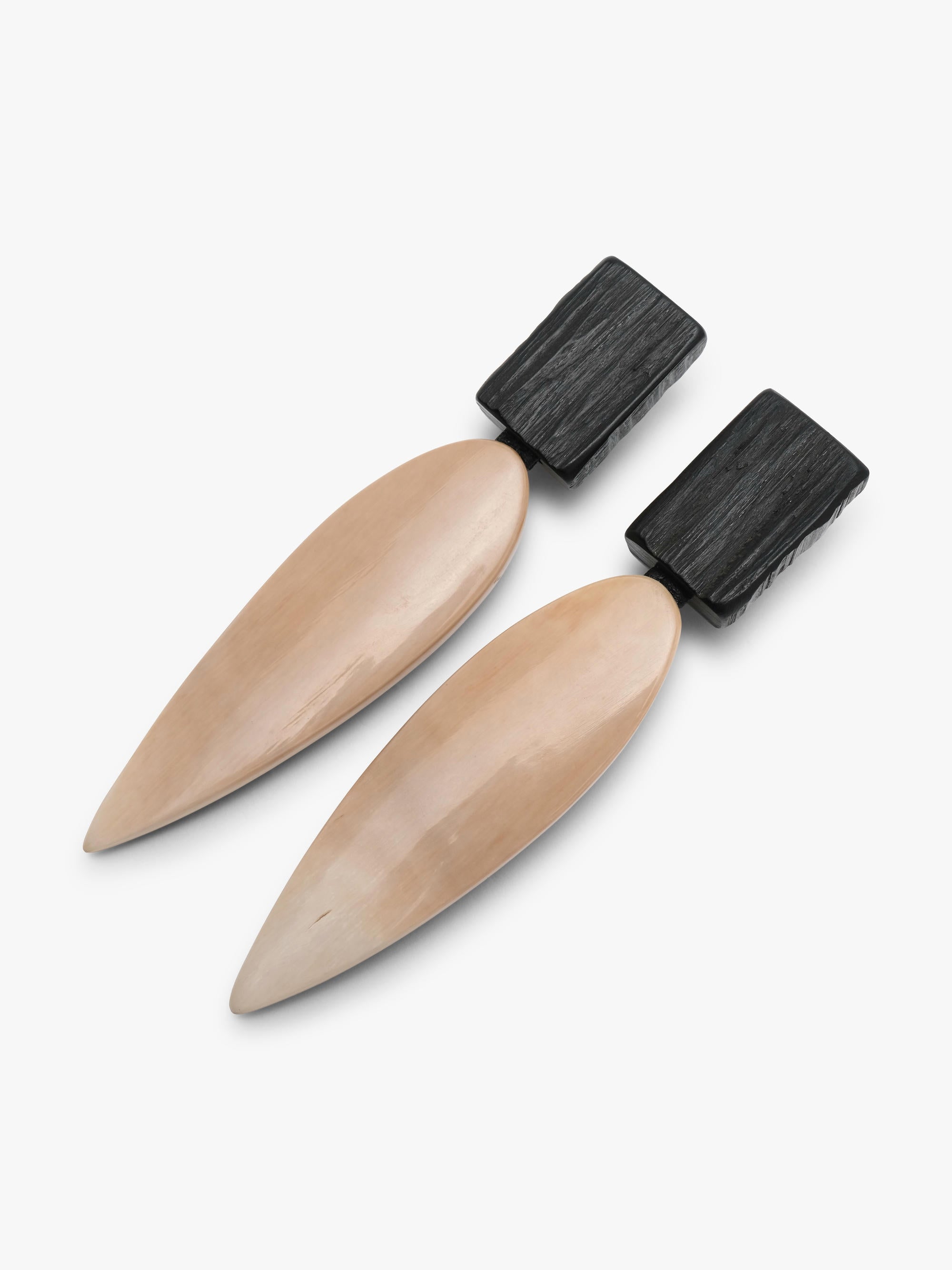A pair of minimalist drop earrings featuring a matte black rectangular top and a teardrop-shaped pendant in a warm champagne beige finish, set against a solid off-white background.