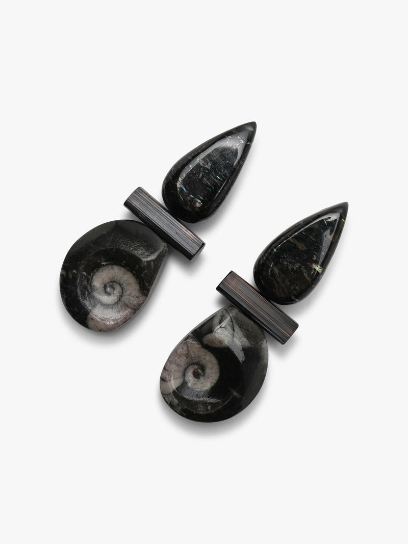 A pair of black teardrop-shaped earrings with a central rectangular bar and a lower drop featuring a polished spiral ammonite shell inlay, shown on a light gray background.