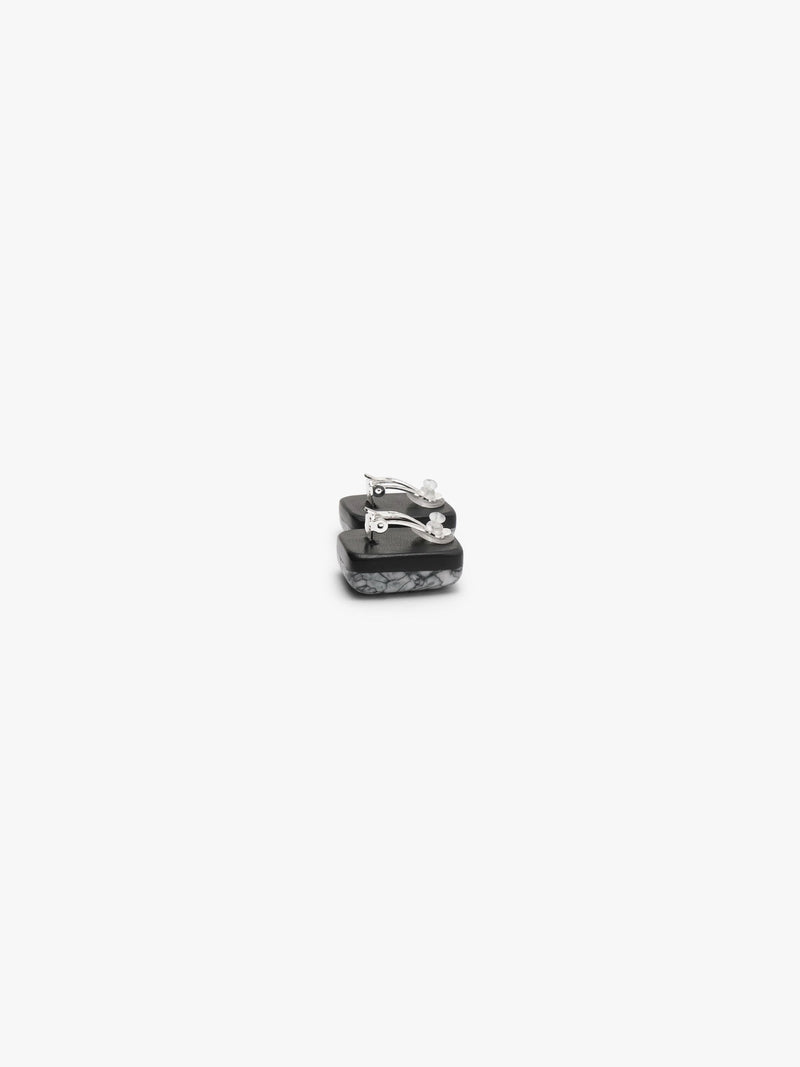 The backside of A pair of small rectangular stud earrings with a textured, marbled gray and black pattern, shown against a solid off-white background.