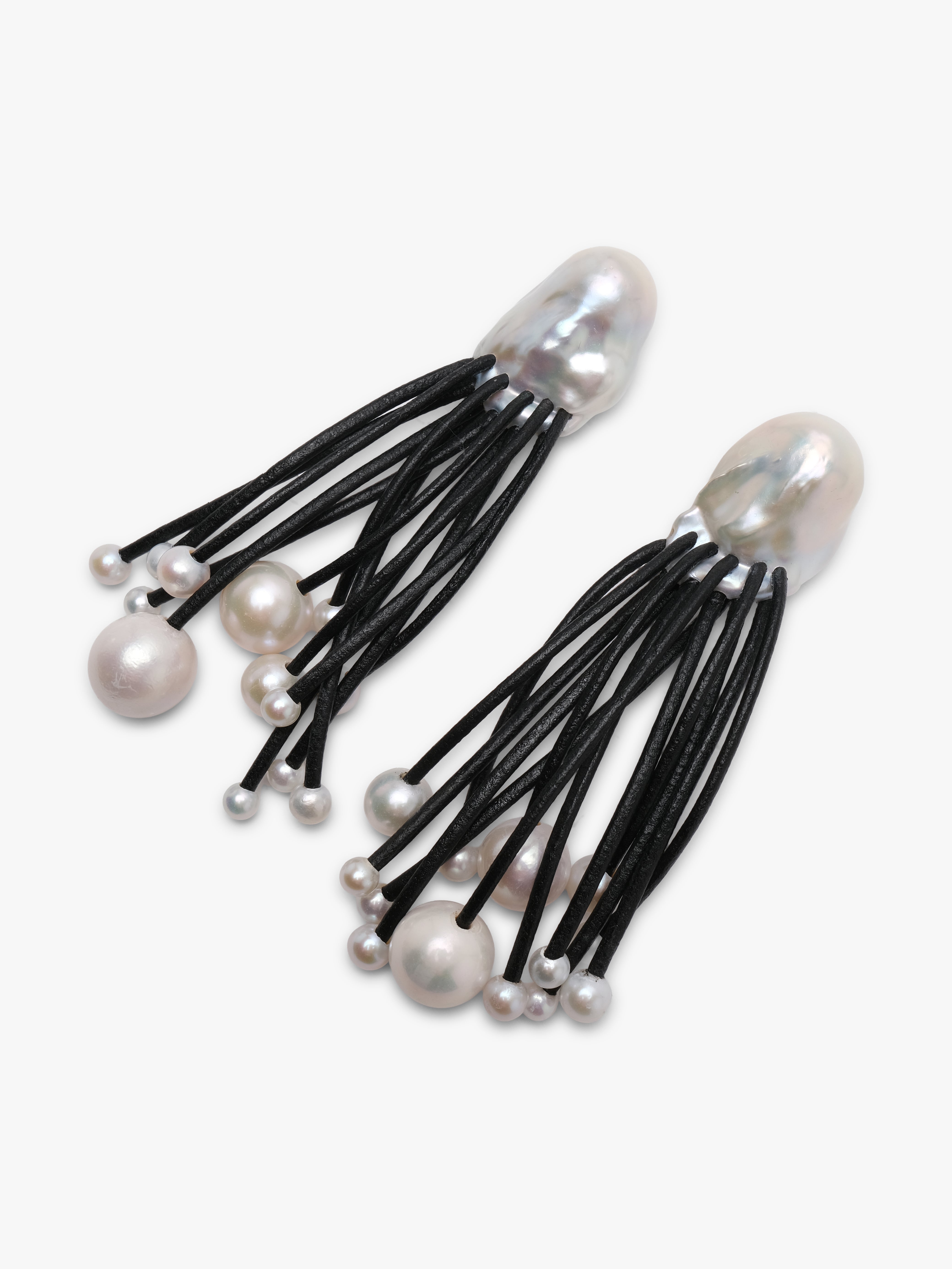 A pair of statement earrings featuring large, irregular white baroque pearls at the top, with multiple thin black cords hanging below, each ending in small white pearl beads, set against a plain light gray background.