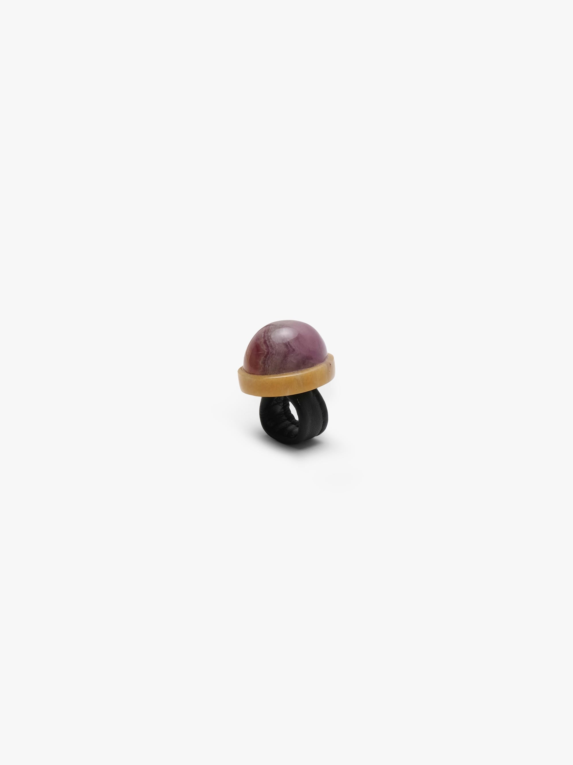 a ring with a dome shaped purple stone set into a creamy golden colored stone and a black leather band. Displayed on a white background.