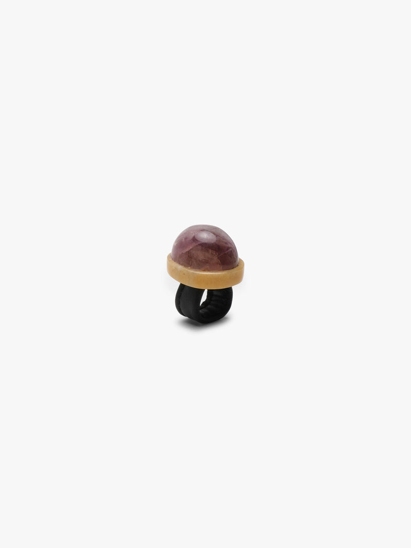 a ring with a dome shaped purple stone set into a creamy golden colored stone and a black leather band. Displayed on a white background.