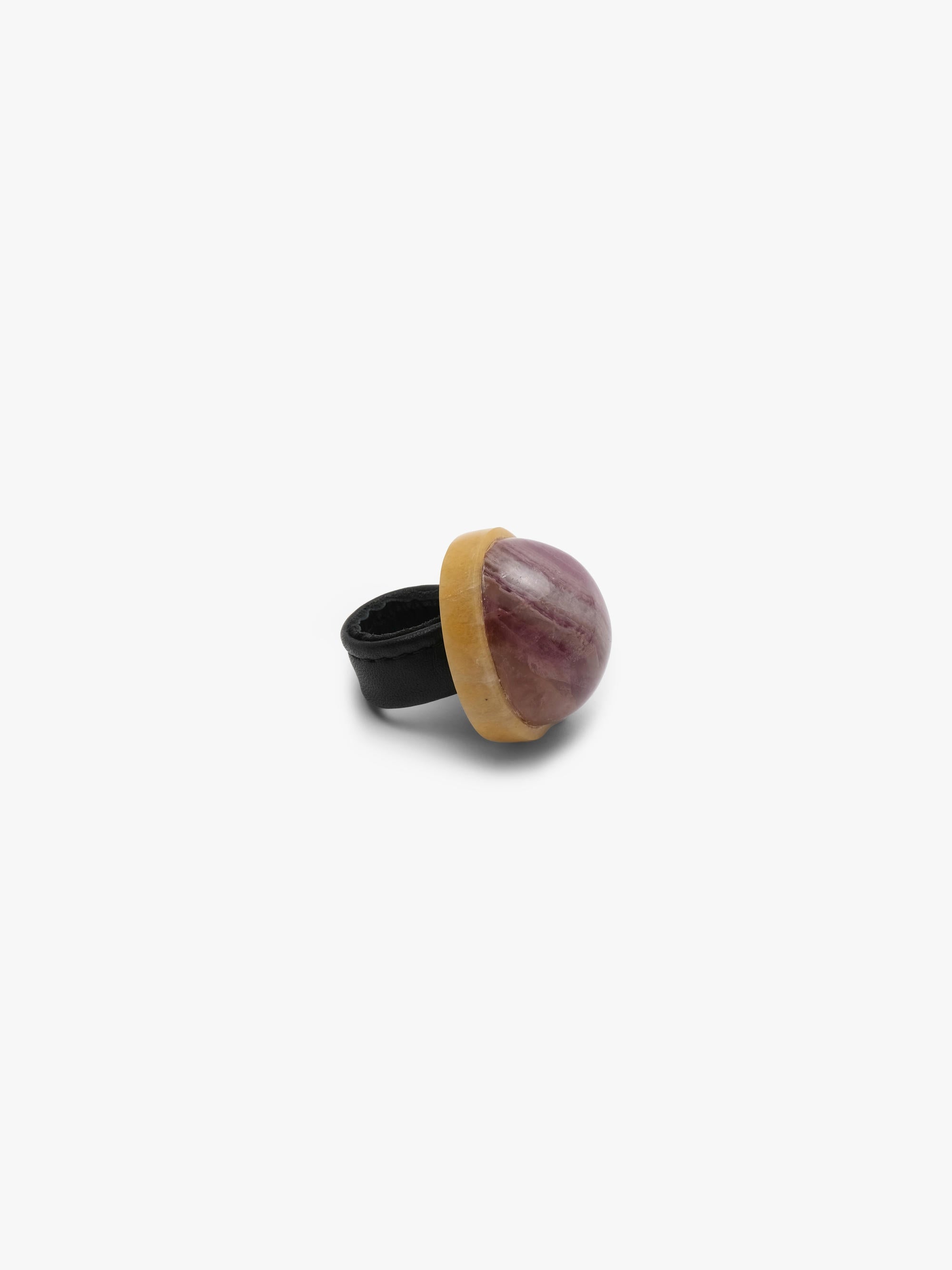 a ring with a dome shaped purple stone set into a creamy golden colored stone and a black leather band. Displayed on a white background.