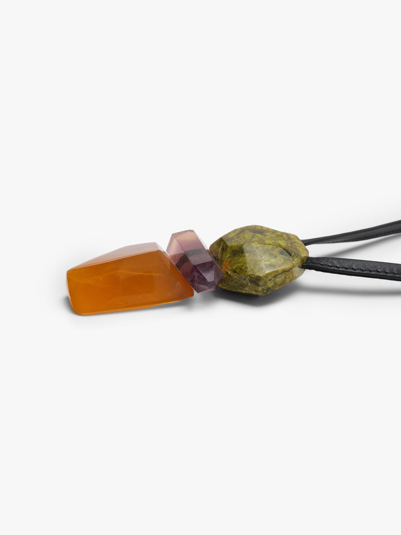 A black cord necklace featuring a stacked crystal pendant with a green stone on top, a pinkish-purple stone in the middle, and a faceted orange stone at the bottom, shown against a plain light gray background.