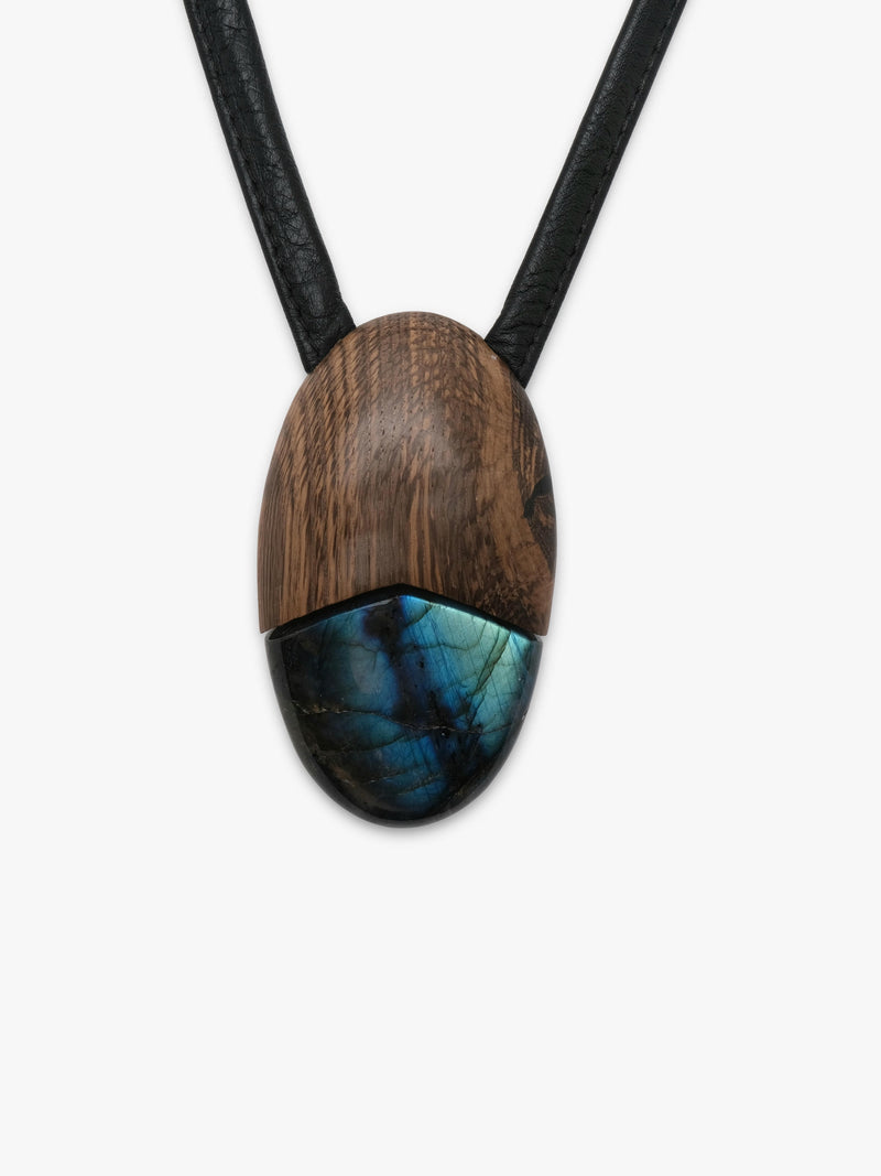 A close-up of a split pendant necklace featuring a dark brown wood top half and a polished blue-green labradorite bottom half, suspended on a black cord against a light gray background.