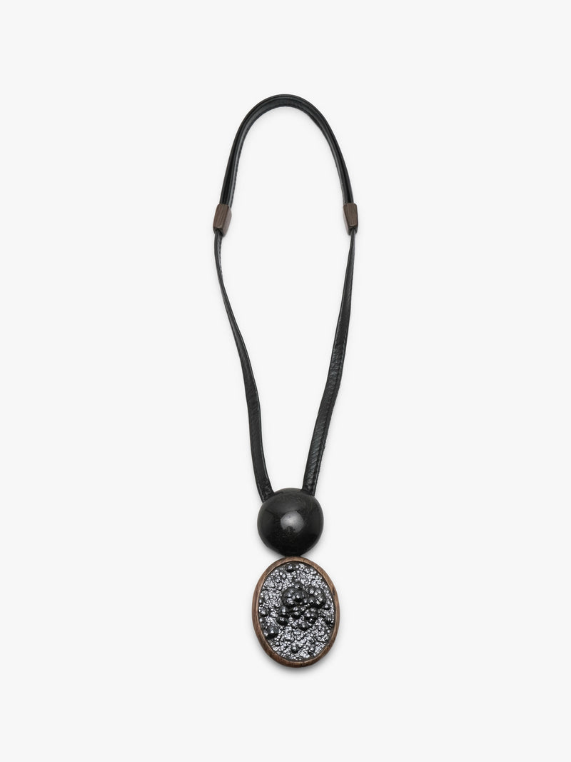 A black leather cord necklace featuring a large round black bead and an oval pendant with a textured black pattern surrounded by a bronze-toned border, shown against a plain white background.