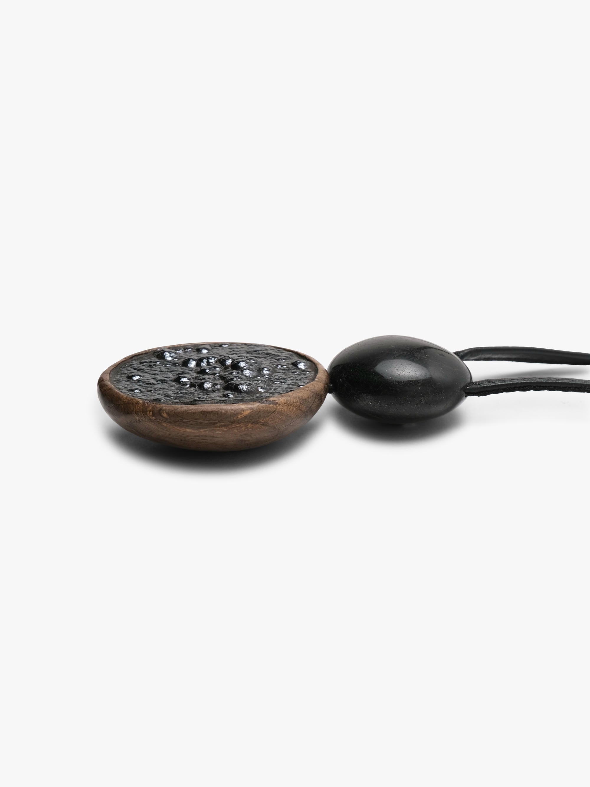 A close-up of a black beaded necklace with a dark brown wooden medallion and a black oval bead on a black cord, set against a plain white background.