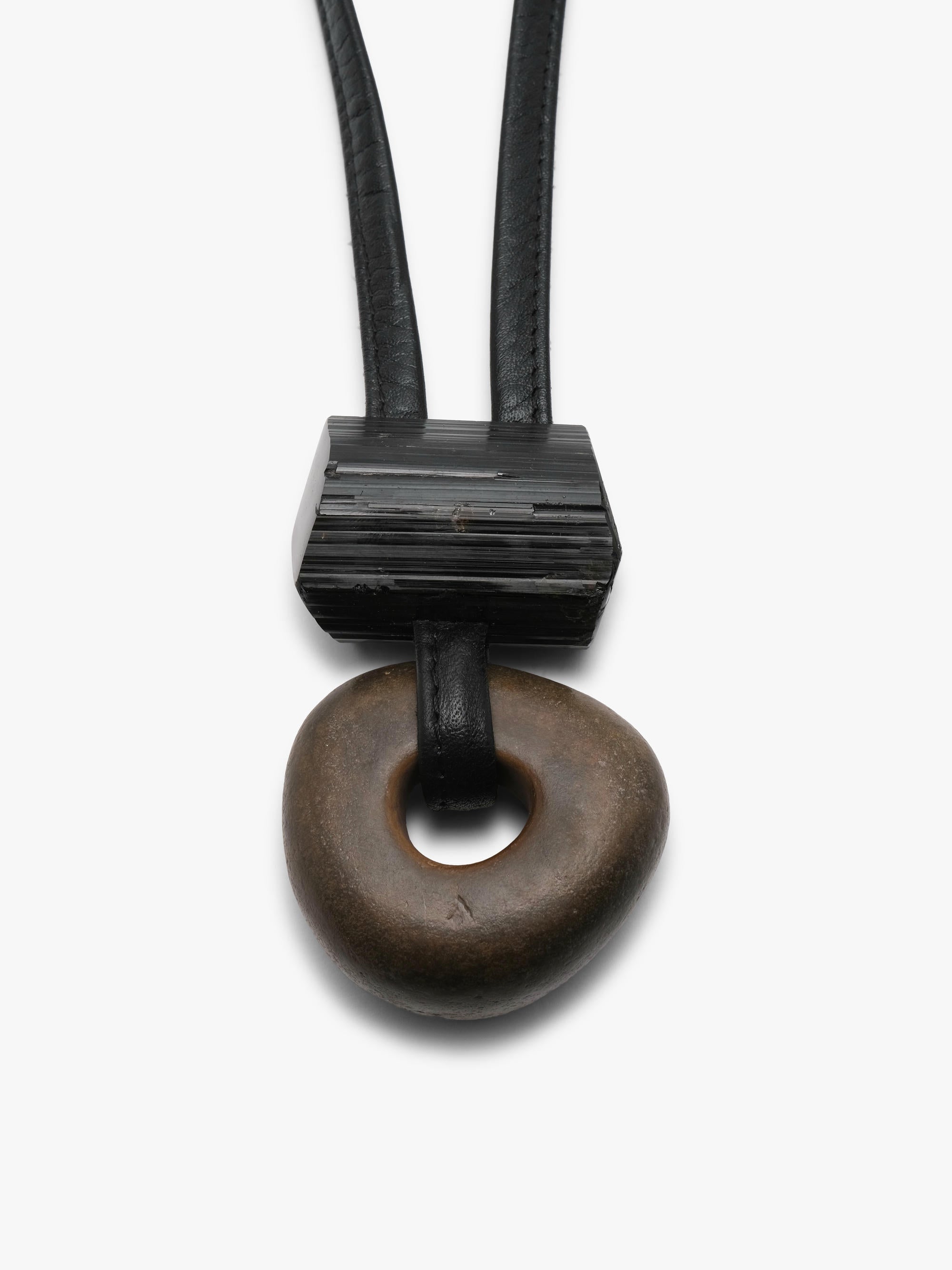 A minimalist black leather and stone pendant necklace featuring a textured black rectangular block and a dark brown circular stone charm, shown against a plain white background.