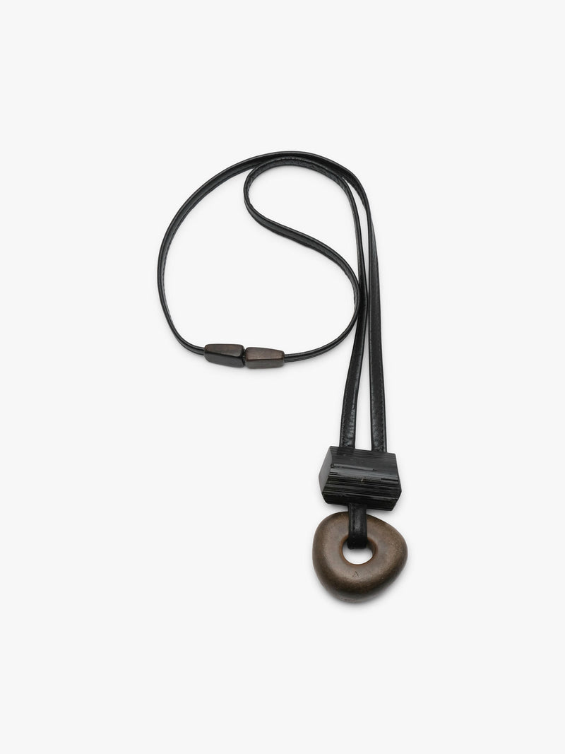 A minimalist black leather and stone pendant necklace featuring a textured black rectangular block and a dark brown circular stone charm, shown against a plain white background.