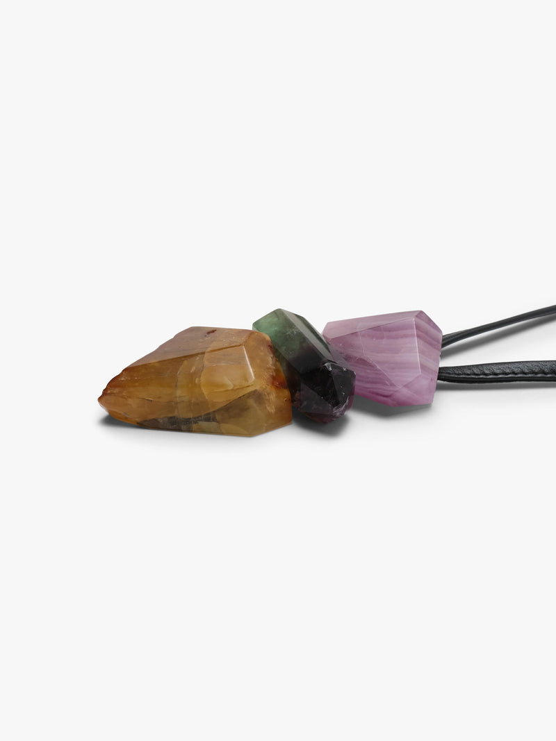 A close-up of a necklace with three faceted crystal pendants in amber-brown, dark green, and lavender-purple, attached to a black cord on a plain white background.