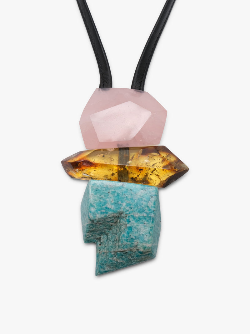 A modern geometric pendant necklace featuring a black elastic cord and a stacked crystal pendant with a pink stone, a yellow-orange stone, and a turquoise stone on a light gray background.