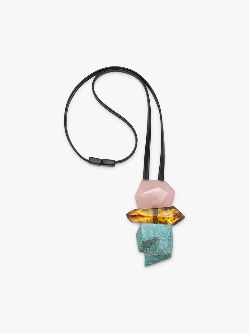 A modern geometric pendant necklace featuring a black elastic cord and a stacked crystal pendant with a pink stone, a yellow-orange stone, and a turquoise stone on a light gray background.
