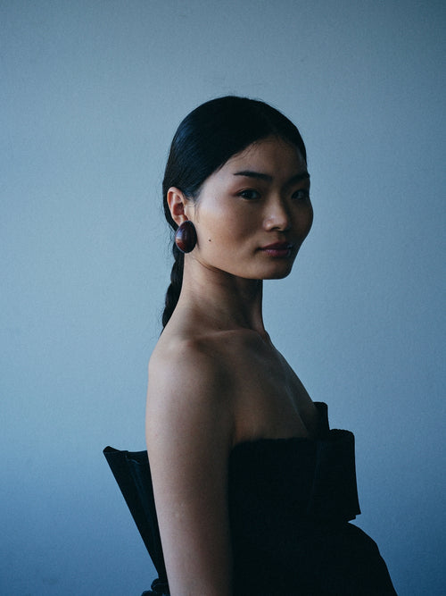 A woman in a black strapless dress wearing large dark circular earrings, photographed against a muted blue-gray background.