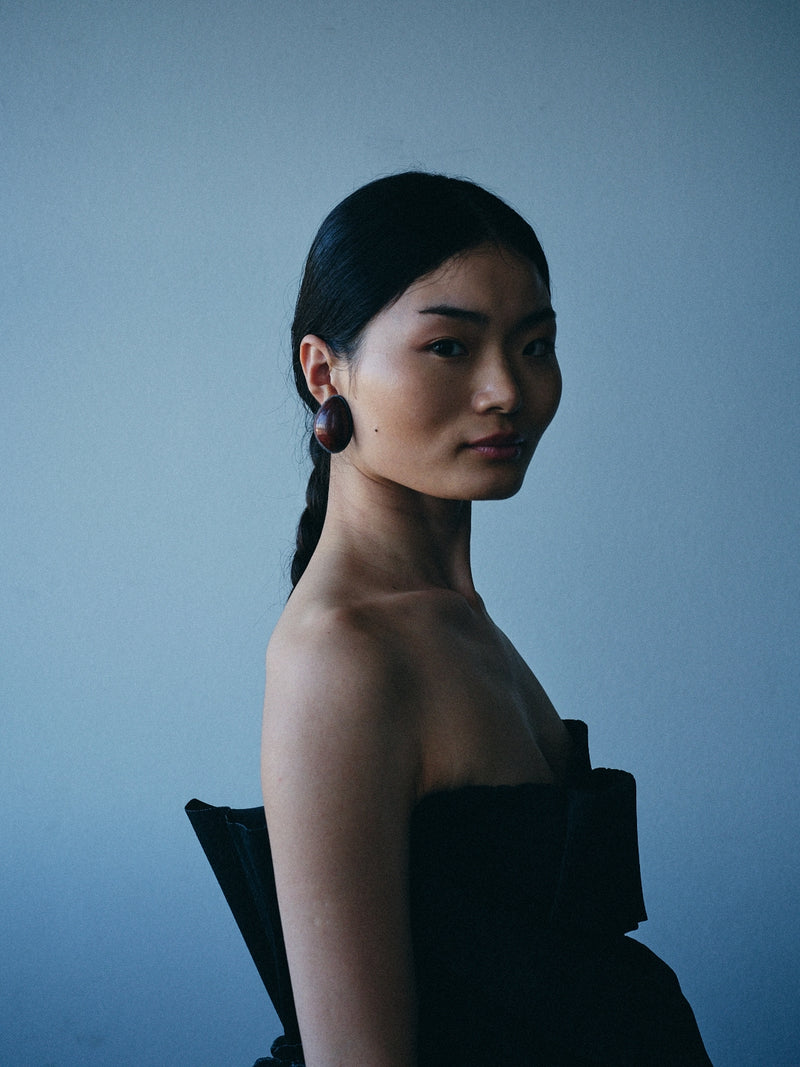 A woman in a black strapless dress wearing large dark circular earrings, photographed against a muted blue-gray background.