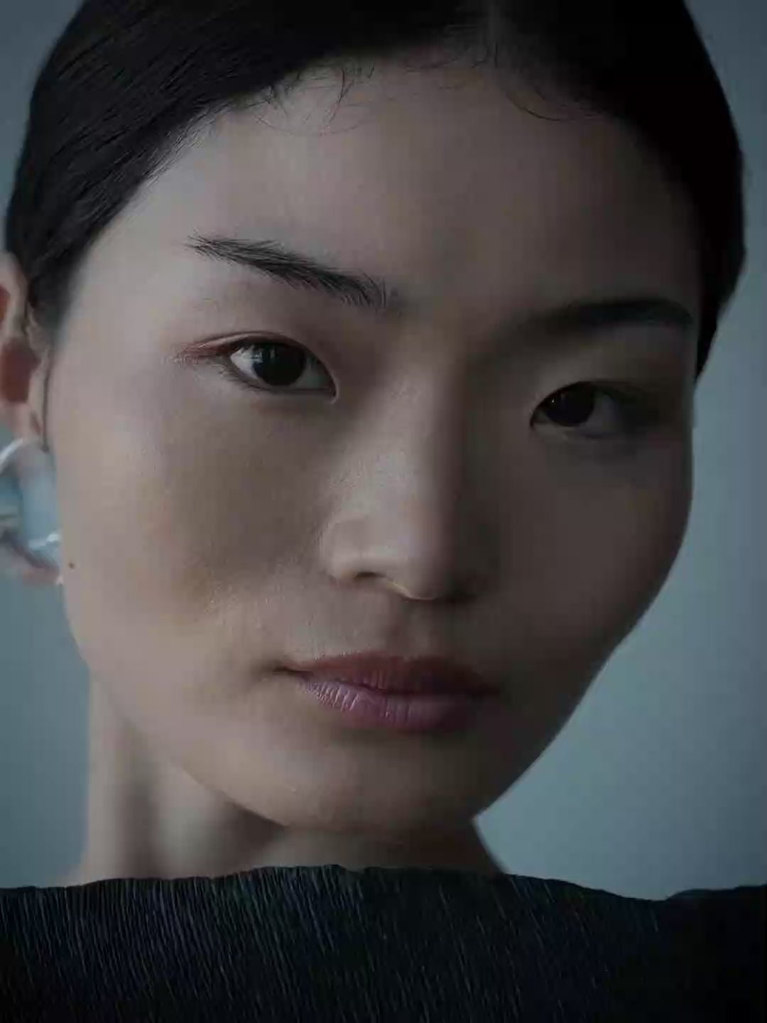 A video of a woman with dark hair tied back, wearing a large, clear, faceted drop-shaped earring against a muted blue-gray background.