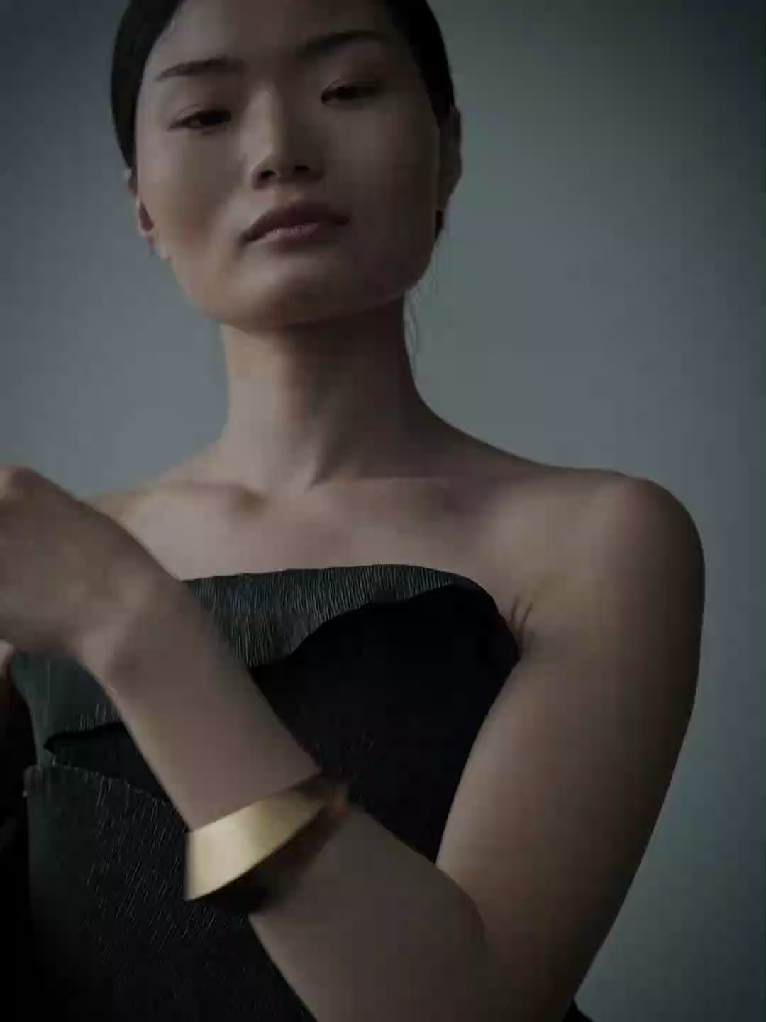 A woman in profile wearing a dark strapless top and a wide gold cuff bracelet, photographed against a muted blue-gray studio background.