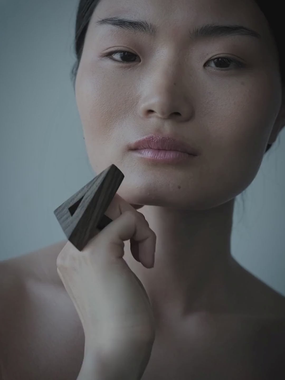 A close-up portrait of a woman with dark hair, wearinga dark geometric triangular ring against her cheek, set against a plain gray background.