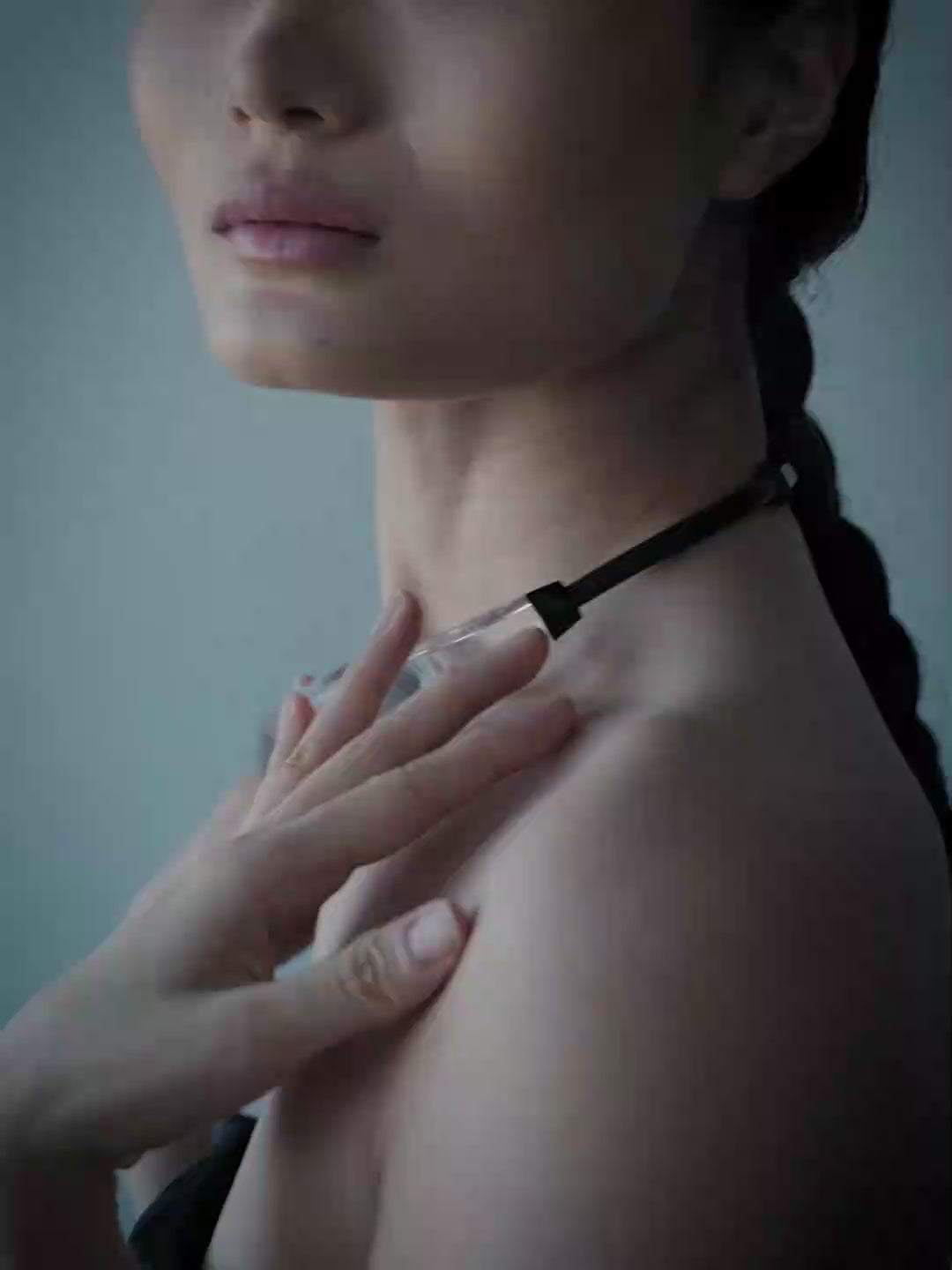 A woman with dark hair pulled back wears a chunky clear faceted geometric collar necklace and a black strapless top, photographed against a solid blue-gray background.